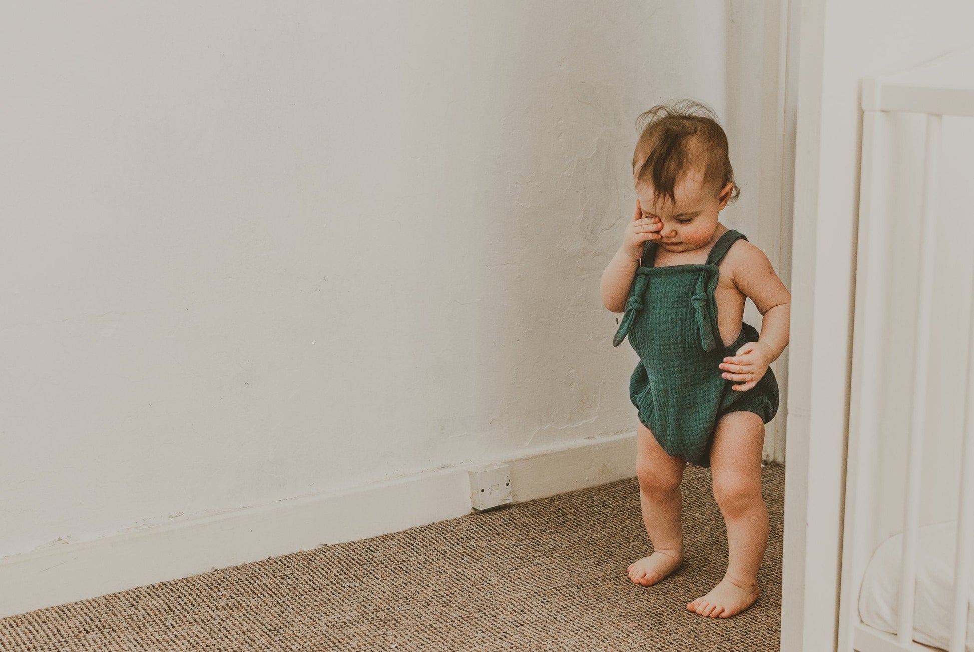 girl walking into a room rubbing her eye wearing the forest green bohemian handmade summer romper with adjustable straps easy to layer up hippy sustainable baby toddler clothing 