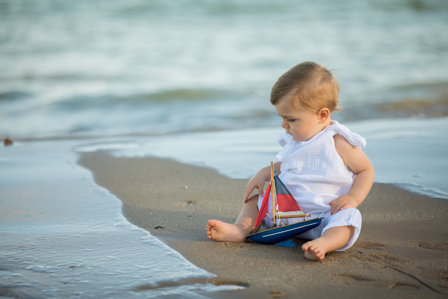 baby boy sat on a beach with the sea in the background playing with a toy boat wearing an adjustable eco friendly romper handmade bohemian hippy style gender neutral