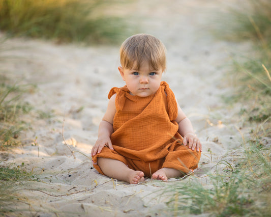 baby boy in a muslin linen handmade romper sat on a beach with adjustable straps bohemian hippy style baby clothing