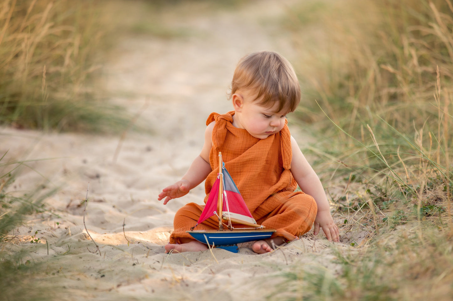 baby boy in a muslin linen handmade romper sat on the sand playing with a toy boat wearing an adjustable straps bohemian hippy style baby clothing unisex