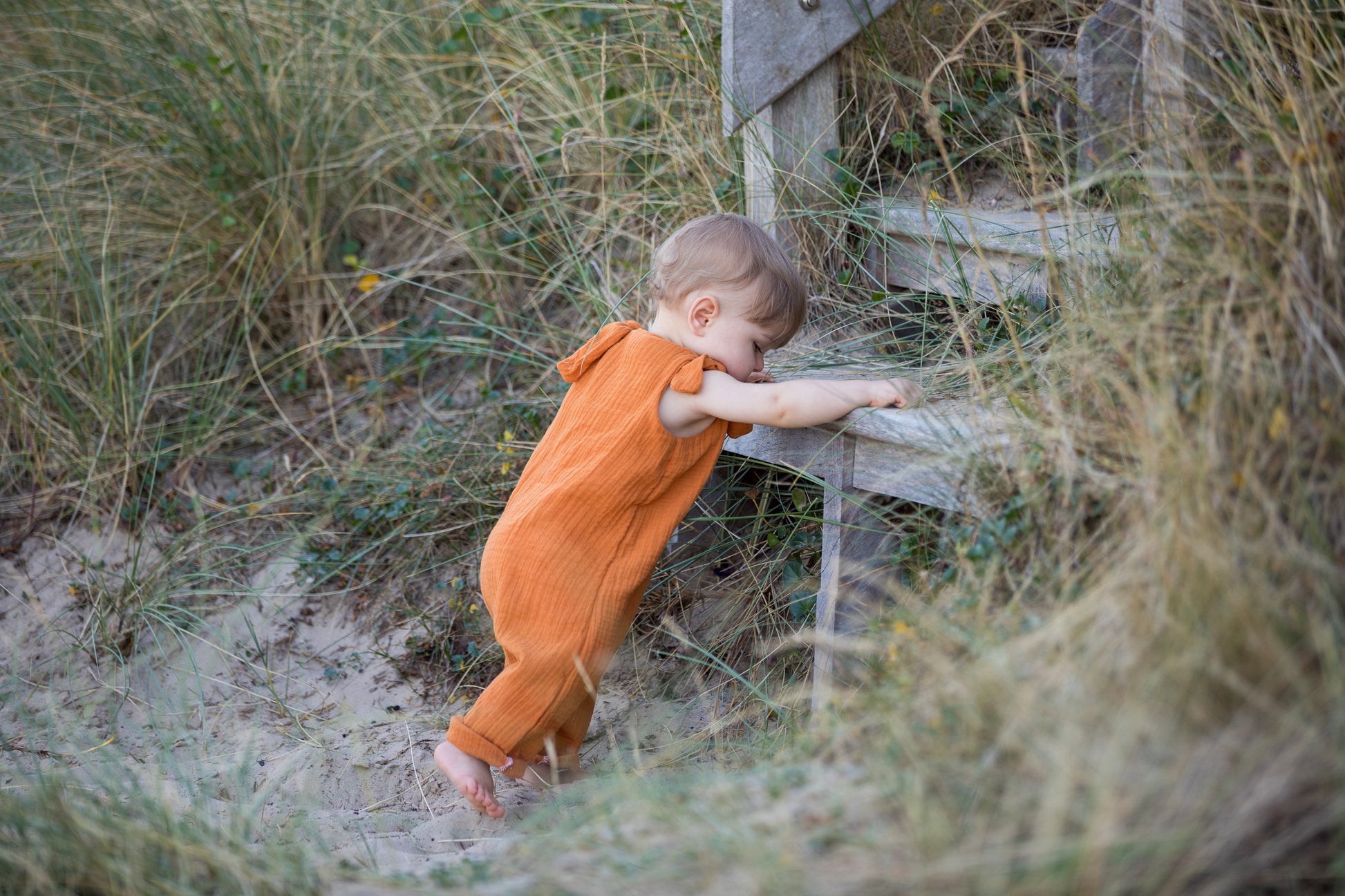 baby boy climbing upstairs on the beach wearing an orange adjustable romper bohemian style unisex