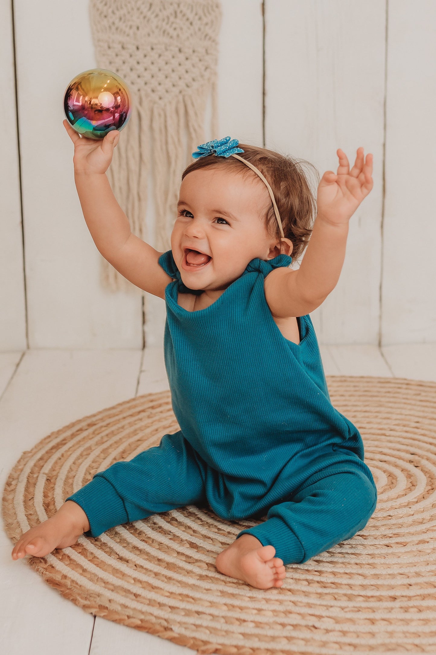 baby girl sits on the bohemian mat playing with a metal ball whilst wearing her handmade teal blue lounge comfy romper adjustable straps cosy winter clothing