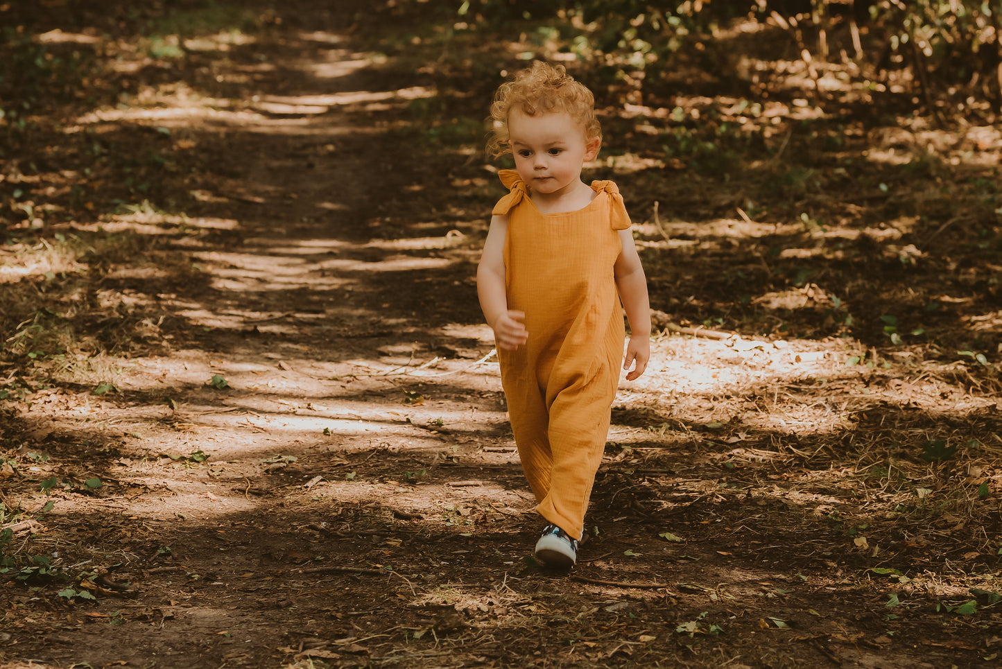 toddler boy walking through a forest in a handmade orange romper with adjustable straps with curly hair bohemian style eco friendly sustainable clothing 