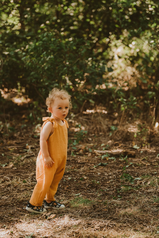 toddler boy stands in forest in orange bohemian romper adjustable straps unisex hippy clothing baby girl curly hair sustainable handmade