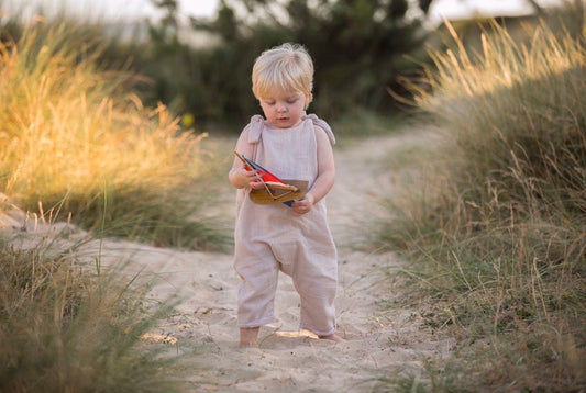 blonde toddler boy stands on the sand holding a toy boat wearing his bohemian hippy style adjustable romper in the colour Natural sustainable baby clothing