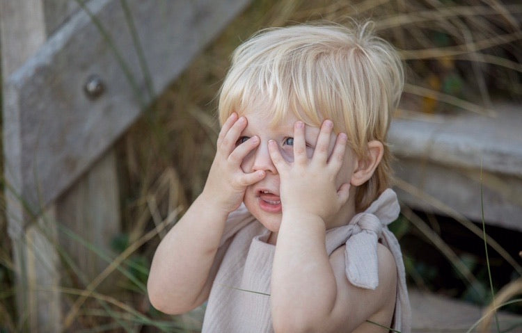 blonde toddler boy sits on the wooden steps on the beach wearing his bohemian hippy style adjustable romper in the colour Natural sustainable baby clothing