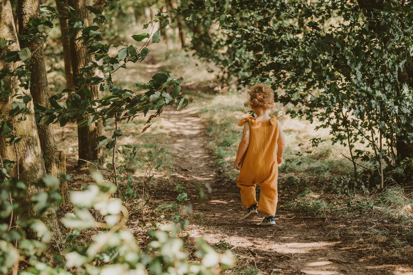 toddler boy walking through a forest in a handmade orange romper with adjustable straps with curly hair bohemian style eco friendly sustainable clothing 