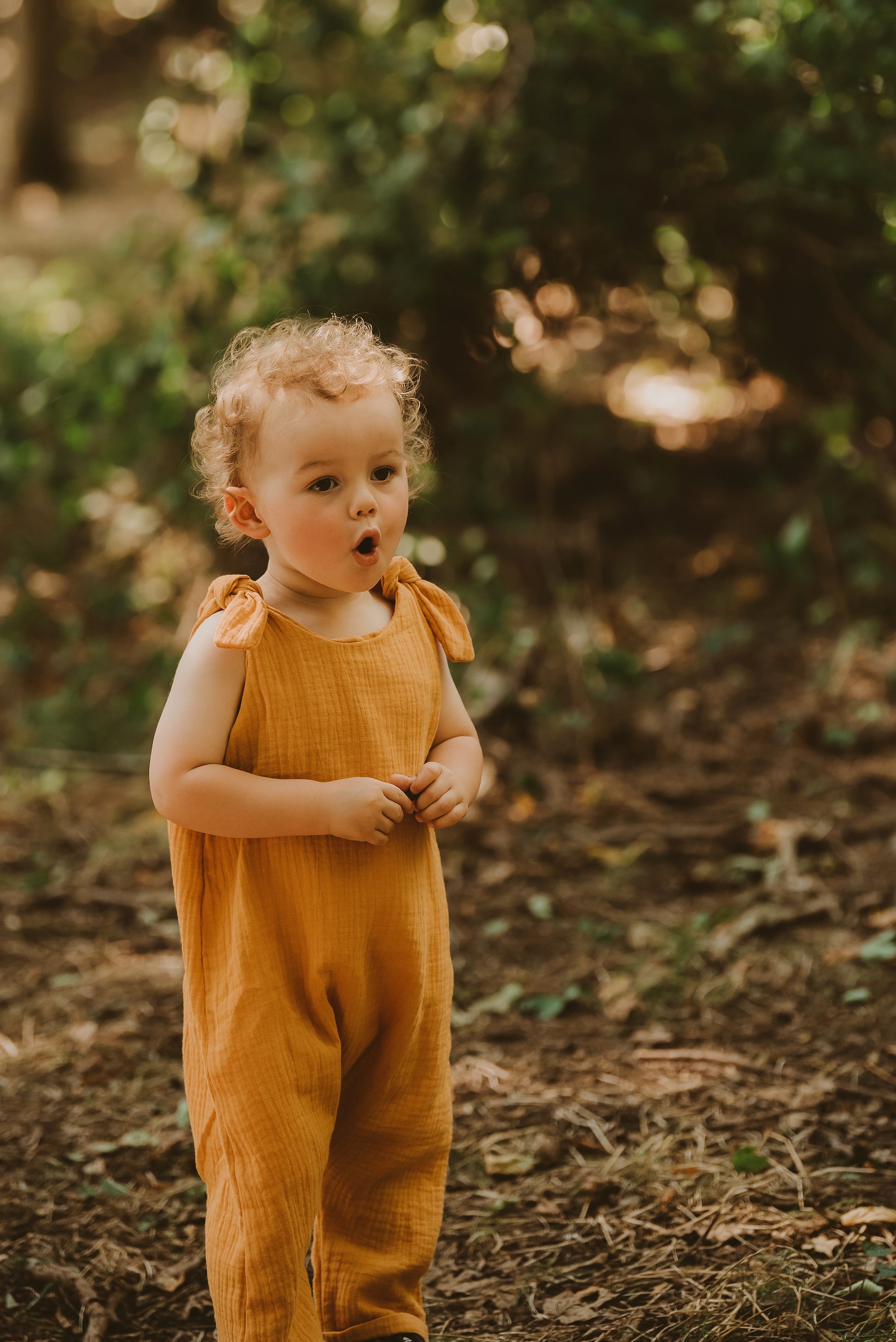 toddler boy stands in forest in orange bohemian romper adjustable straps unisex hippy clothing baby girl curly hair sustainable handmade