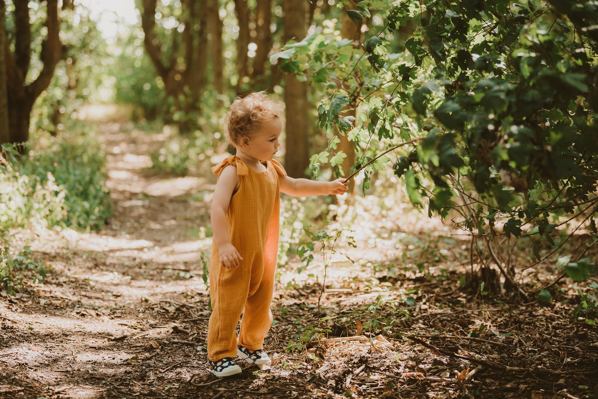 toddler boy stands in forest in orange bohemian romper adjustable straps unisex hippy clothing baby girl curly hair sustainable handmade