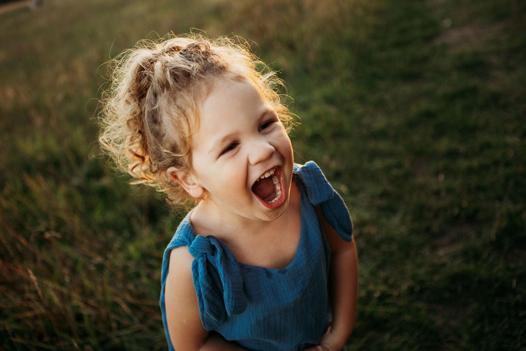 toddler boy stood in a meadow sunset behind laughing wearing handmade bohemian romper with adjustable straps in blue curly hair hippy style clothing unisex eco friendly sustainable 