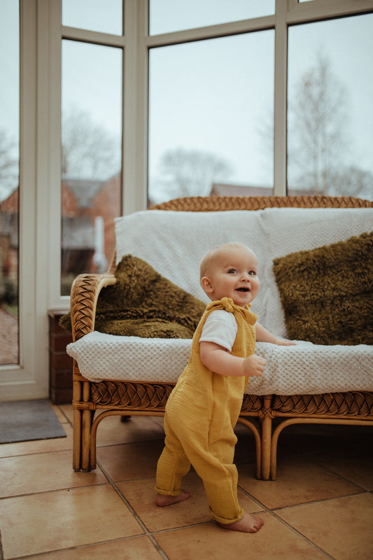 toddler boy girl stood in mustard romper adjustable straps in summer conservatory wicker furniture surrounding unisex eco friendly sustainable gender free clothing bohemian baby girl hippy style handmade