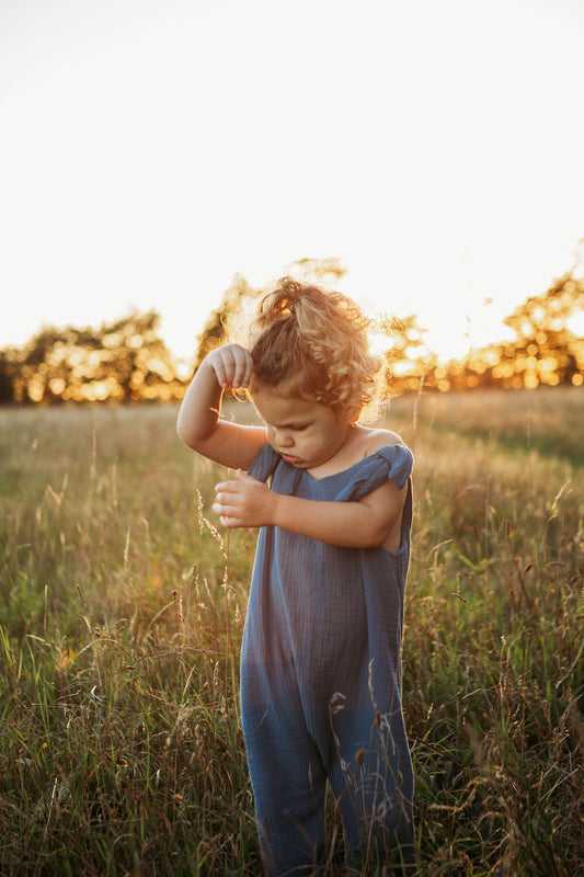 toddler boy stood in a meadow sunset behind wearing handmade bohemian romper with adjustable straps in blue curly hair hippy style clothing unisex eco friendly sustainable 